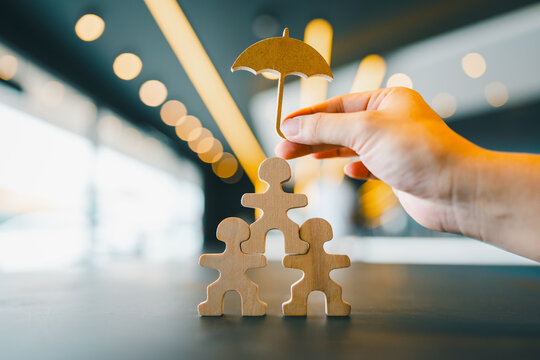 Hand holding umbrella icon over wooden human figures in pyramid shape, symbolizing insurance protection, family care, team safety, risk management, and employee welfare concept.
