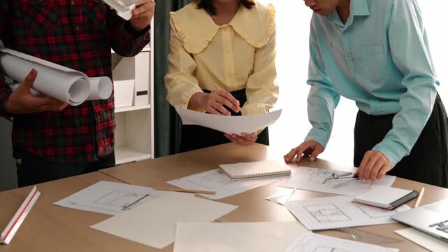Architect and safety engineer discussion about building blueprint displaying on laptop on table with architectural document and wooden block scatter around at modern office. Closeup. Delineation.