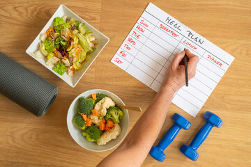 Healthy eating and nutrition control with weekly meal planner, fresh vegetables, salad bowl, yoga mat and dumbbells for balanced diet and fitness routine at home.