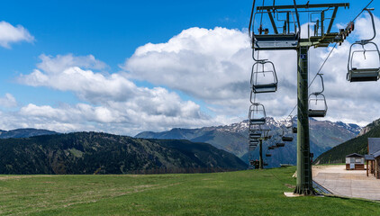 a ski lift in its off-season repose, with empty chairs hanging over a grassy slope, set against a backdrop of mountain ranges under a sky with fluffy clouds