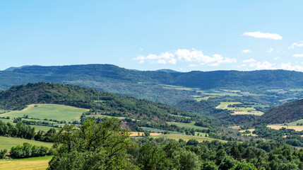Rolling hills covered in lush green fields stretch across the landscape in the Pyrenees. The sunlit valleys, dotted with patches of trees, create a picturesque scene under a clear blue sky.