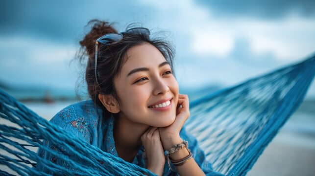 Happy young Asian woman on a hammock at the seaside, smiling and enjoying leisure, blue sky with clouds, tropical vacation atmosphere, wide empty space for website or advertising text.