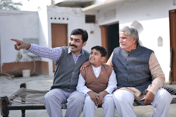 Fototapeta premium Concept of Third Generations. Indian happy young father pointing finger to son and his grand father sitting in village. Happy Parents Day.
