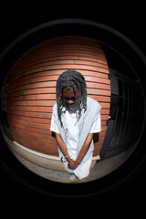 Fish eye view of Black young man standing outdoors with head bowed, hands clasped in front, wearing casual clothing, dreadlocks visible, brick wall background, not looking at camera