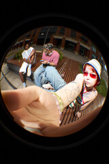 Young Caucasian woman with tattoos taking selfie using smartphone with fisheye lens while sitting on bench outdoors, Black and Caucasian man sitting in background