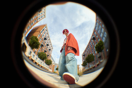 Caucasian young adult woman with short pink hair standing outdoors in urban setting, wearing oversized clothing, looking down at camera with confident expression, fisheye lens effect