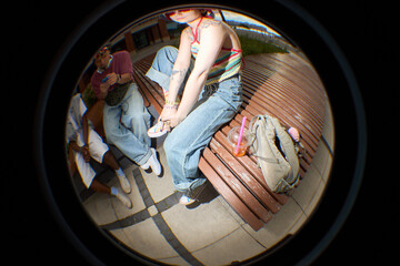 Fisheye lens view of young Caucasian woman sitting on bench adjusting shoe, showing tattoos on arm, next to Black man and friend, casual outdoor setting, backpack and drink nearby
