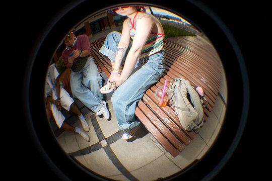 Fisheye lens view of young Caucasian woman sitting on bench adjusting shoe, showing tattoos on arm, next to Black man and friend, casual outdoor setting, backpack and drink nearby