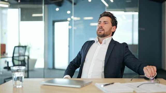 Satisfied professional businessman in a dark suit and white shirt at an office desk closes laptop, removes glasses, and leans back, feeling happy and content in a modern office.