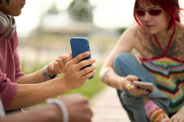 Fototapeta premium Young Asian man showing smartphone screen to Caucasian woman with red hair and tattoos, both sitting outdoors and interacting with mobile devices on summer day