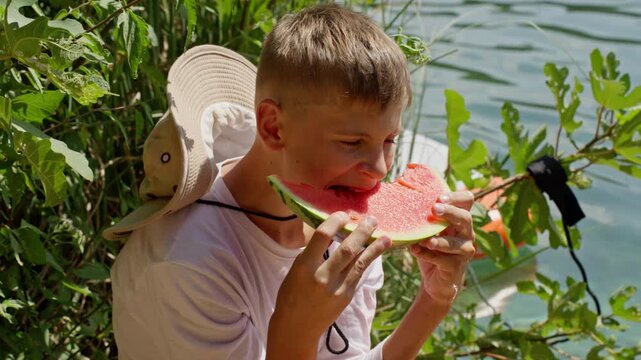Happy teenage boy with hat sitting by lake on sunny summer day, hungrily biting juicy red slice of fresh watermelon during picnic or hike in beautiful green nature