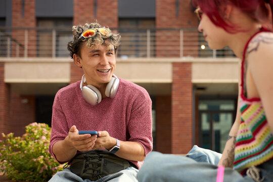 Young Caucasian man with curly hair and headphones around neck, smiling and holding smartphone, while talking to young female friend with red hair and visible tattoos outdoors - Powered by Adobe