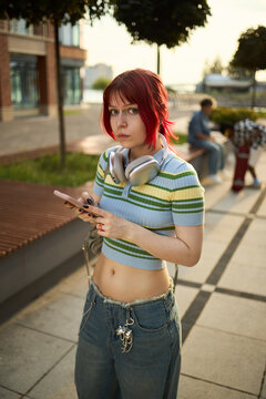Fototapeta Portrait of Caucasian teenager girl with red hair standing outdoors holding smartphone wearing headphones around neck looking at camera with serious expression urban background