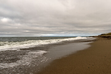 Waves roll across the sandy shoreline beneath a gray overcast sky, with foamy surf spreading along the coast, capturing the raw beauty of a natural seascape. Sea of Okhotsk Sakhalin Island