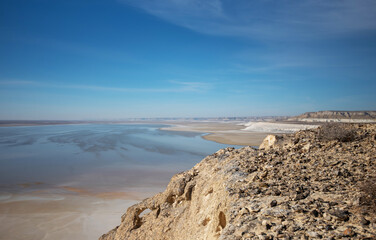 A panoramic view of the Boszhira Valley on the Ustyurt Plateau in Kazakhstan. The rugged terrain with rocky cliffs meets the vast salt flats, extending to the horizon under a deep blue sky.