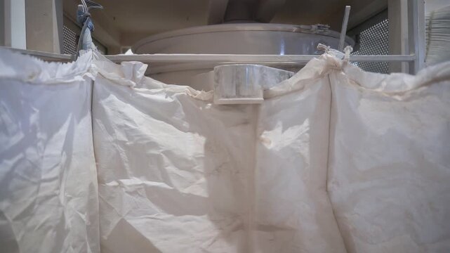 Filling big plastic bag with dry dust like material from metal pipe in plant workshop. Chemical components packaging into large sack at manufacturing facility