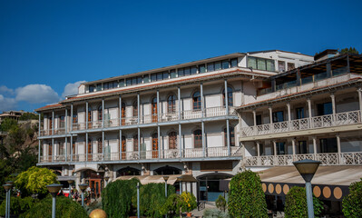 Fototapeta premium A traditional Georgian building with arched windows and wooden balconies in Tbilisi. Surrounded by greenery and sunny weather, the structure features elegant architecture and historical charm.
