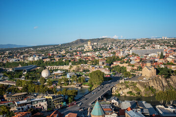 aerial view of Tbilisi, Georgia, showcasing the Kura River, ancient churches, and blend of historic and modern architecture under a clear blue sky, with greenery and scenic landscapes around the city © eskstock