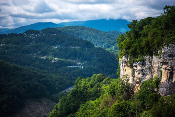 a dramatic landscape featuring a rugged cliff adorned with greenery, overlooking a valley with scattered houses amidst rolling, forest-covered hills under a moody sky.