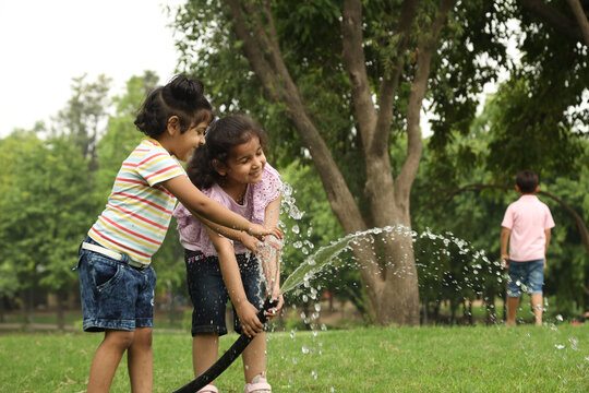Indian happy and cute kid girls playing and splashing water pipe in garden. Background of green grass watering theme concepts