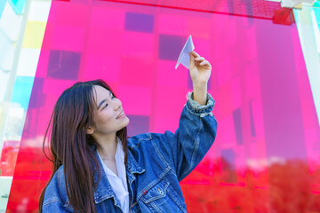 Young woman smiling, holding a paper airplane in hand, getting ready to launch it against a vibrant, colorful background