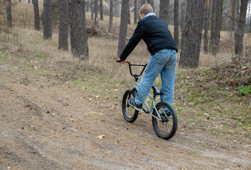 Rear view of a young man riding a BMX bike on a natural dirt path through a pine forest. This...