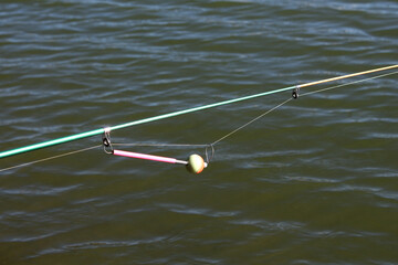 Fishing rod float, fish bite detector. Fishing rod with green blank and thin line extended over water, equipped with a bright strike indicator featuring pink stick and round float for bite detection.