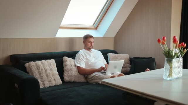 A man is sitting on a comfortable sofa at home, using a laptop for remote work or browsing. Natural daylight comes through a skylight window, creating a cozy and modern home atmosphere. Fresh tulips