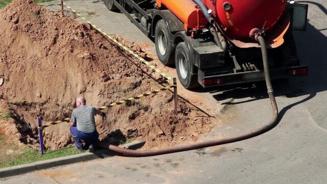 pumping tank against the background of a pit with a fence. Concept of liquidation of an emergency breakdown in a water supply system, copy space for text. A male worker operates a hose to pump out a w