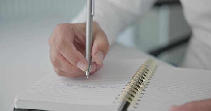 Close-up of hands - a man in a shirt writes with a pen and takes notes in a notepad on a metal spring on an office desk