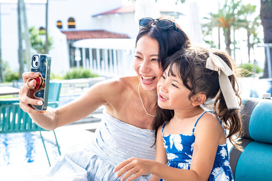 Japanese mother and daughter taking a close up selfie together at a resort poolside café during summer vacation smiling and posing with tropical palm trees and holiday atmosphere - Powered by Adobe