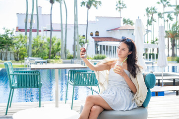 A Japanese woman sitting by a resort swimming pool holding a cold drink and taking a selfie with her smartphone while enjoying a relaxing summer holiday