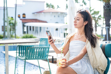 A Japanese woman relaxing at a resort poolside holding a refreshing drink in one hand and looking at her smartphone while enjoying a calm vacation atmosphere