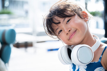 A young Japanese girl smiling playfully while wearing white headphones around her neck at a resort poolside area on a sunny summer day with tropical background and relaxed vacation atmosphere