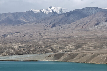 Aerial view of the stark, arid landscape meeting the serene turquoise waters of Baisha Lake beneath snow-capped mountains, Baisha Lake, Xinjiang, China.