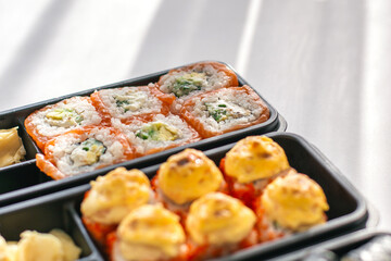 Close-up of assorted sushi rolls in black plastic container on white table in sunlight. Fresh salmon rolls and baked rolls with cheese topping, served with wasabi and pickled ginger