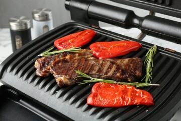 Electric grill with beef steaks, bell peppers and rosemary on white table, closeup
