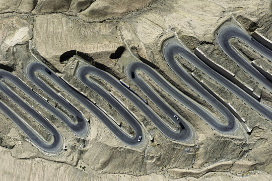 Aerial view of a snaking asphalt road cutting through the stark, arid landscape of Panlong Ancient Road, a ribbon of dark contrast against the pale, textured earth, Panlong Ancient Road, China.