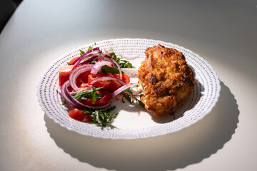 On a decorative Plate is a piece of fried chicken, alongside a tomato and red onion salad. It sits on a white table under natural light, possibly in a kitchen or dining room.