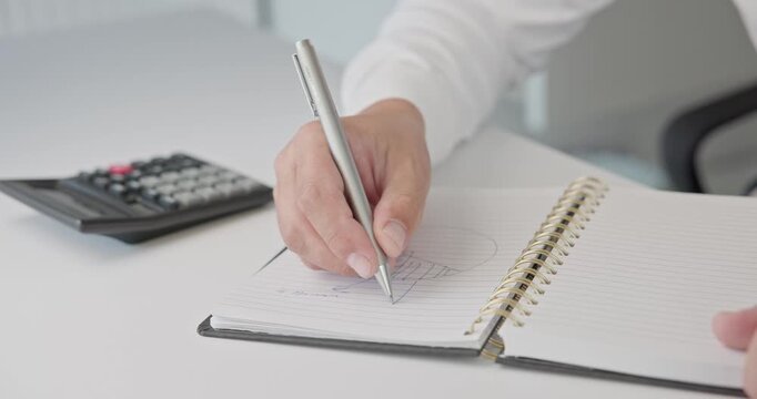 Close-up of hands - a man in a shirt draws a diagram and takes notes with a pen in a notepad on a metal spring on an office desk