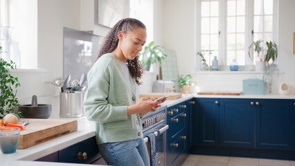 Obraz premium Teenage Girl At Home In Kitchen Connecting With Friends On Social Media Using Mobile Phone