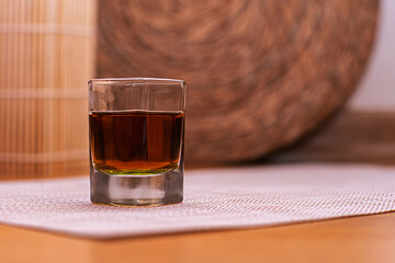 The glass half-filled with a dark brown drink. Beverage placed on a textured mat over a wooden surface. The background features warm tones of woven and bamboo textures. Giving a cozy and  rustic feel.