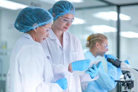 A senior scientist mentors a junior researcher, pointing at results on a digital tablet as they collaborate on an experiment in a bustling, modern scientific laboratory environment.