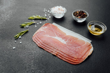 Pack of sliced prosciutto ham with salt, pepper and herbs on black background