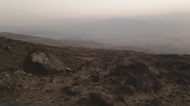 Ararat base camp views. Rocky volcanic slope at dusk above wide valley, south Turkey. Sparse steppe, Kurdish homeland concept, endurance trekking mood, remote wilderness, long trail toward horizon.