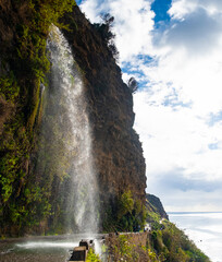Cascata dos Anjos waterfall flows dramatically over a coastal road in Madeira, Portugal. Visitors drive through the cascading water, creating a unique and memorable experience