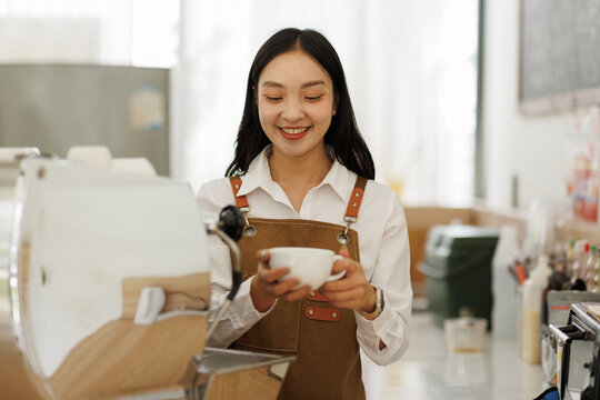 Young asian woman barista wearing apron using professional coffee grinder and managing orders in modern cafe