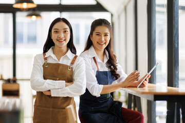 Two Asian female waitress wearing apron using digital tablet taking orders from customer in cafe, SME entrepreneur seller business concept
