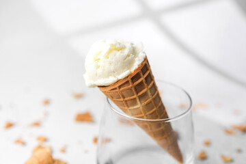 Closeup of waffle cone with creamy vanilla ice cream resting in clear glass on bright background with crumbs