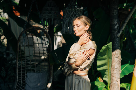 A woman in an elegant outfit stands with her eyes closed in the rays of the sun on a terrace in a tropical garden.